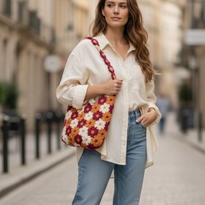 May include: A colorful crochet tote bag with a floral pattern in shades of white, orange, and burgundy. The bag has a matching strap and is carried over the shoulder. The background is a blurred city street.
