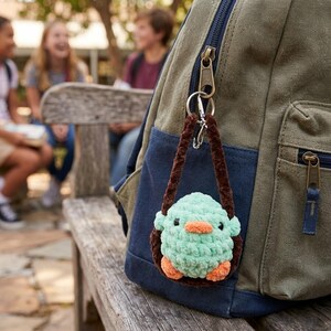 May include: A handmade, mint green crochet keychain shaped like a small bird, with orange feet and beak. The keychain is attached to a brown loop and hangs from a green and blue backpack. The background shows a wooden bench and children.