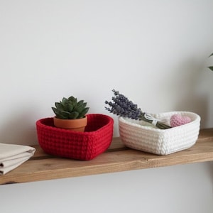 May include: Two heart-shaped crochet baskets, one red and one white, sit on a wooden shelf. The red basket holds a small potted succulent. The white basket contains lavender and a small pink heart. A folded beige cloth is on the shelf.