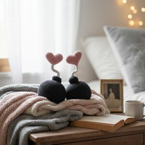 May include: Two black crochet bombs with pink heart-shaped tops sit on a plush blanket. The bombs have white stems and are placed on a wooden table. A framed photo, a mug, and an open book are also on the table.
