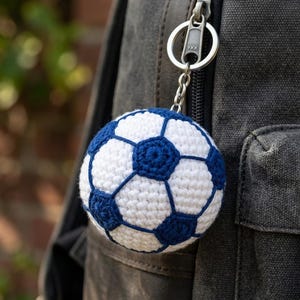 Puede incluir: Un llavero de balón de fútbol hecho a mano con ganchillo. La pelota presenta un diseño clásico con paneles blancos y azules, sujeto a un llavero y cadena plateados. El llavero está colgado de una mochila gris.