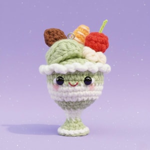 Puede incluir: Un helado de ganchillo con una cara sonriente, con helado verde, una cereza y otros ingredientes. El helado está en una copa blanca y verde con un borde con volantes, sobre un fondo púrpura.