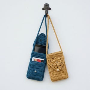 K&ouml;nnte beinhalten: Zwei handgefertigte geh&auml;kelte Handytaschen h&auml;ngen an einem dekorativen Haken. Eine ist tiefblau, mit einer Tasche f&uuml;r ein Telefon und Karten. Die andere ist senfgelb, mit einem Blumendesign und Perlenakzent. Beide haben lange Tr&auml;ger.