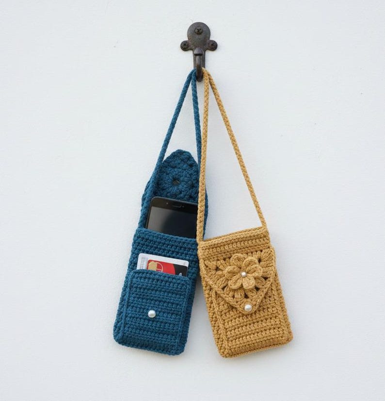 K&ouml;nnte beinhalten: Zwei handgefertigte geh&auml;kelte Handytaschen h&auml;ngen an einem dekorativen Haken. Eine ist tiefblau, mit einer Tasche f&uuml;r ein Telefon und Karten. Die andere ist senfgelb, mit einem Blumendesign und Perlenakzent. Beide haben lange Tr&auml;ger.