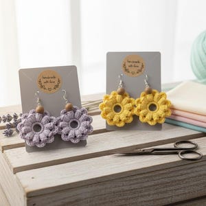 May include: Two pairs of handmade crochet flower earrings. One pair is lavender, the other is yellow. Each earring has a wooden bead and silver-coloured hook. The earrings are displayed on grey cards with the text "handmade with love".