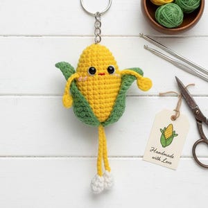 May include: A handmade crochet corn keychain with a yellow body, green husk, and dangling yellow legs with white feet. The keychain has a silver ring and a tag that says "Handmade with Love."
