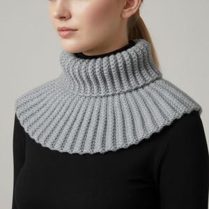 Könnte beinhalten: Ein grauer Strick-Rollkragenwärmer. Der Halswärmer ist gerippt und hat einen gerüschten Rand. Die Person auf dem Bild trägt ein schwarzes Langarmshirt. Der Rollkragenwärmer ist ein Winteraccessoire.