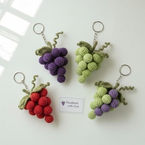 Pode incluir: Quatro chaveiros de uvas de crochê feitos à mão em vermelho, verde e roxo. Cada chaveiro apresenta um cacho de uvas com folhas verdes e um chaveiro prateado. Um pequeno cartão com o texto "Handmade with Love" também é visível.
