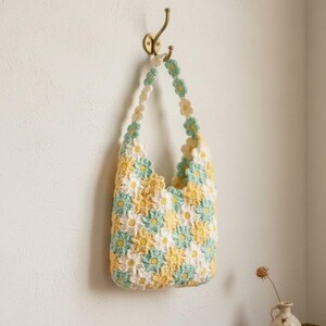 May include: A handmade crochet bag with a daisy pattern in yellow, white, and light blue. The bag is hanging on a brass hook against a white wall. The strap is made of daisy-shaped crochet flowers.