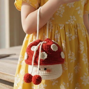 May include: A red and white crochet mushroom-shaped bag with a drawstring closure and a white strap. The bag features white polka dots and two red pom-poms. The bag is being held by a person wearing a yellow dress.