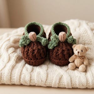 Puede incluir: Un par de patucos de bebé de ganchillo marrón hechos a mano con detalles de hojas verdes y un detalle de bellota rosa. Un pequeño osito de peluche beige con un lazo está sentado junto a los patucos sobre una manta de punto de cable color crema.