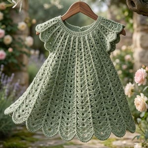 Puede incluir: Un vestido de ganchillo verde salvia con dobladillo festoneado y mangas cortas cuelga de una percha de madera. El vestido presenta un patrón calado detallado y se muestra sobre un fondo borroso al aire libre con flores.