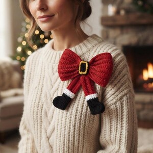 Peut inclure: Un pull en tricot torsadé couleur crème orné d'un grand nœud au crochet sur le thème du Père Noël. Le nœud est rouge vif avec une boucle de ceinture noire et jaune au centre, et deux jambes pendantes avec des revers blancs et des bottes noires.