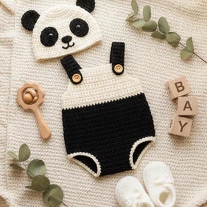 Puede incluir: Un conjunto de ropa de bebé hecho a mano, que incluye un gorro de panda, un pelele y patucos. El pelele es negro y crema, con detalles de botones de madera. También hay una sonaja de madera y bloques que deletrean "BAY". El conjunto está sobre una manta crema.