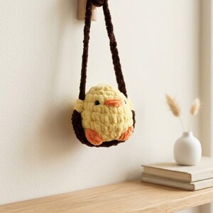 May include: A handmade yellow crochet chick-shaped bag with brown straps. The chick has orange feet and a beak, and black eyes. The bag is hanging on a wooden hook. A small white vase with dried flowers and a stack of books are on a wooden shelf.