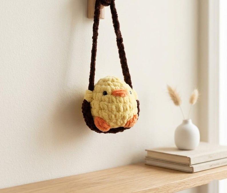 May include: A handmade yellow crochet chick-shaped bag with brown straps. The chick has orange feet and a beak, and black eyes. The bag is hanging on a wooden hook. A small white vase with dried flowers and a stack of books are on a wooden shelf.