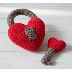 May include: A red crocheted heart-shaped lock with a gray handle and keyhole detail, next to a red heart-shaped key with a gray handle. The handmade items are on a white wooden surface.