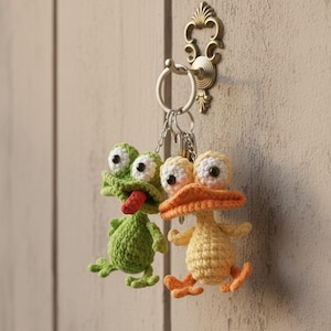 May include: Two handmade crochet keychains: a green frog with a red tongue and a yellow duck with an orange beak and feet. Both have large, googly eyes. They hang from a metal keyring attached to a decorative brass hook on a wooden surface.