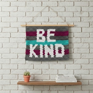 Puede incluir: Un tapiz de pared de ganchillo con las palabras "BE KIND" en letras blancas. El fondo es gris con rayas horizontales turquesas y burdeos. El tapiz está suspendido de una barra de madera con un cordón natural.