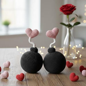 Puede incluir: Dos bombas de ganchillo negras con corazones rosas y tallos blancos. Pequeños corazones de ganchillo rojos y rosas están dispersos. Una rosa roja en un jarrón de cristal está en el fondo. Sobre una superficie de madera.