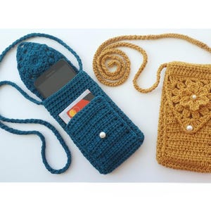 May include: Two handmade crochet phone pouches. One is teal blue with a pocket and a white pearl button. The other is mustard yellow with a floral design and two white pearl accents. Both have long, matching straps.