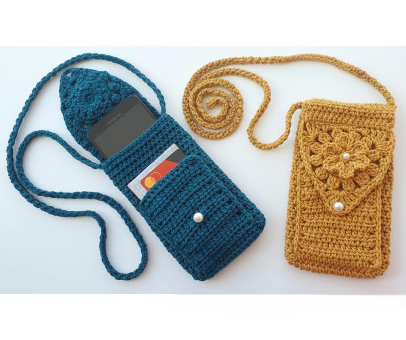 K&ouml;nnte beinhalten: Zwei handgefertigte geh&auml;kelte Handytaschen. Eine ist t&uuml;rkisblau mit einer Tasche und einem wei&szlig;en Perlmuttknopf. Die andere ist senfgelb mit einem Blumendesign und zwei wei&szlig;en Perlen. Beide haben lange, passende Tr&auml;ger.