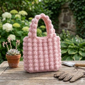 Può includere: Una borsa tote all'uncinetto rosa chiaro fatta a mano con un motivo a bolle strutturato. La borsa ha un manico arrotondato e poggia su un tavolo di legno. Sono visibili anche una piccola pianta grassa in vaso e guanti da giardinaggio.