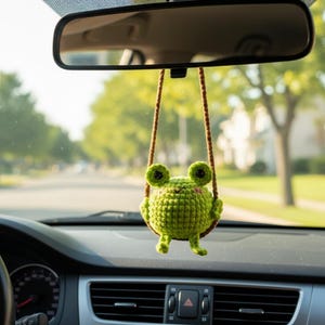 May include: A green crocheted frog car charm hanging from a brown string, suspended in front of a car's dashboard and rearview mirror. The frog has large eyes and a smiling face, adding a whimsical touch to the vehicle's interior.