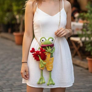 以下が含まれることがあります： 赤いカニのアクセサリーが付いた、気まぐれなカギ針編みのカエル型ハンドバッグ。カエルは大きな目、ピンクの口、黄色のオーバーオールが特徴です。バッグはゴールドチェーンから吊り下げられており、楽しくて個性的なアクセサリーを提供しています。