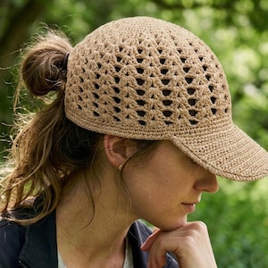 May include: A beige crochet baseball cap with a peak. The cap features an open weave pattern. The cap is worn by a person with brown hair tied back. The cap is made from a soft, natural fibre.