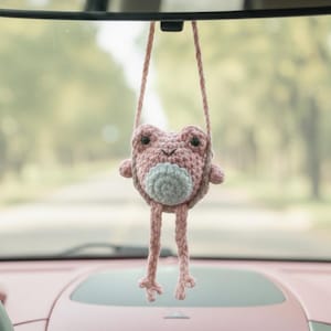 May include: A handmade, crocheted frog car charm in shades of pink and gray, hanging from a string. The frog has a round body, small arms, and long legs. The charm is suspended in front of a car's windshield.