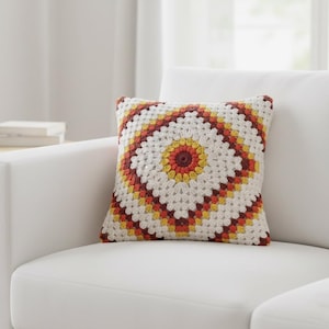 May include: A square crochet pillow with a geometric design in white, yellow, orange, and burgundy. The pillow is placed on a white sofa. The crochet pattern features a central circular design surrounded by diamond shapes.
