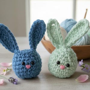 Peut inclure: Deux figurines de lapin au crochet faites à la main. L'une est bleue, l'autre est vert clair. Les deux ont des yeux noirs et des nez roses en forme de cœur. Les lapins ont de longues oreilles et sont posés sur une surface claire. De la laine et des crochets sont en arrière-plan.