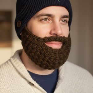 May include: A dark navy blue knit beanie with a green leaf design and a brown crocheted beard. The beard is a dark brown color and is attached to the beanie. The person is wearing a white sweater.