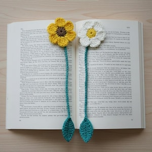 Puede incluir: Dos marcadores de libros hechos a mano con flores de ganchillo sobre un libro abierto. Uno presenta una flor amarilla con centro marrón, el otro una flor blanca con centro amarillo. Ambos tienen tallos y hojas de color verde azulado.