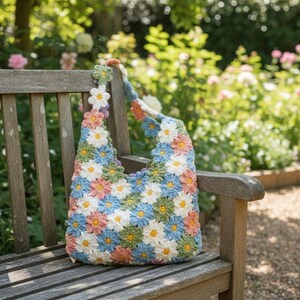 May include: A handmade crochet tote bag featuring a daisy pattern in white, blue, pink, green, and yellow. The bag is sitting on a wooden bench in a garden setting. The strap is also adorned with crochet daisies.
