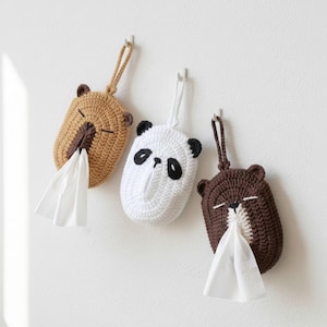 May include: Three crochet tissue box covers shaped like animals: a bear, a panda, and a bear. The bear is tan, the panda is white with black accents, and the other bear is brown. Each has a loop for hanging and a slot for tissue dispensing.