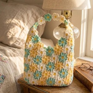 May include: A handmade crochet bag featuring a floral pattern in yellow, white, and light blue. The bag has a rounded shape with a handle adorned with daisy-like flowers. It sits on a wooden nightstand next to a lamp and a book.