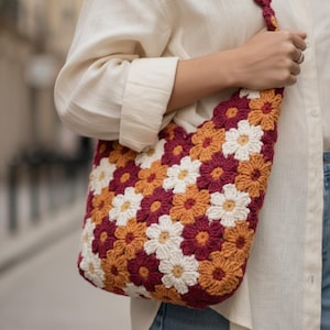以下が含まれることがあります： 白、バーガンディ、オレンジ色の花柄が特徴の、かぎ針編みのショルダーバッグ。バッグにはお揃いのストラップが付いています。クリーム色のシャツを着た人がバッグを持っています。