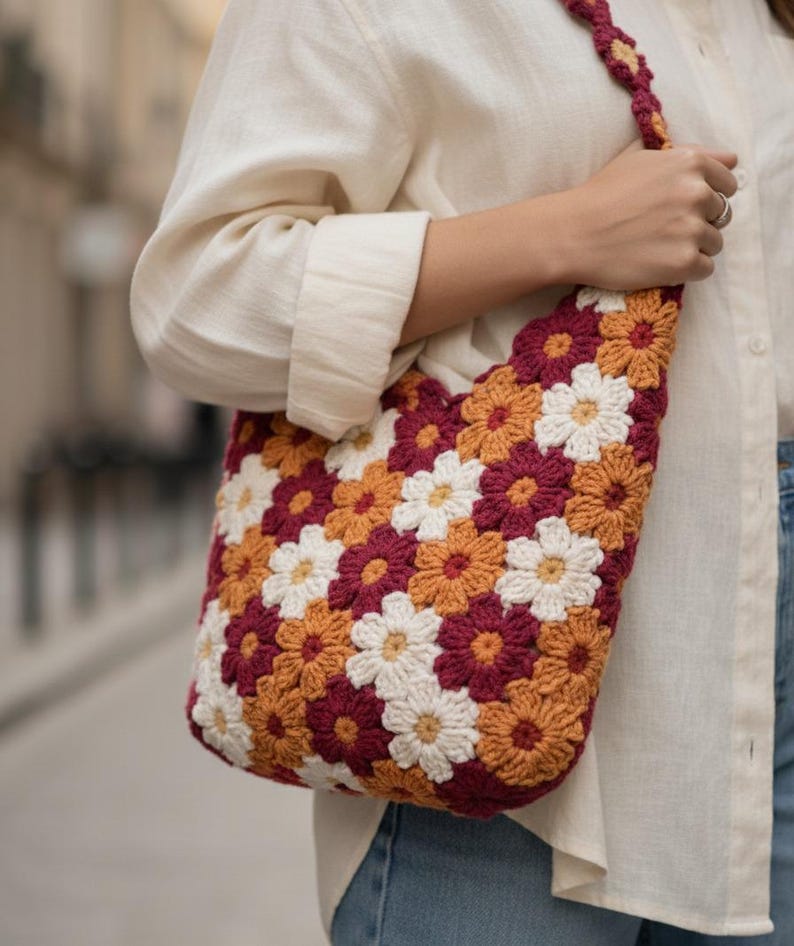 May include: A crocheted shoulder bag featuring a floral pattern in white, burgundy, and orange. The bag has a matching strap and is held by a person wearing a cream-colored shirt.