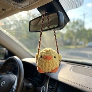 May include: A yellow crocheted chick car charm hanging from a car's rearview mirror. The chick has orange feet and a beak, with brown string straps. The interior of the car is visible, including the steering wheel and dashboard.