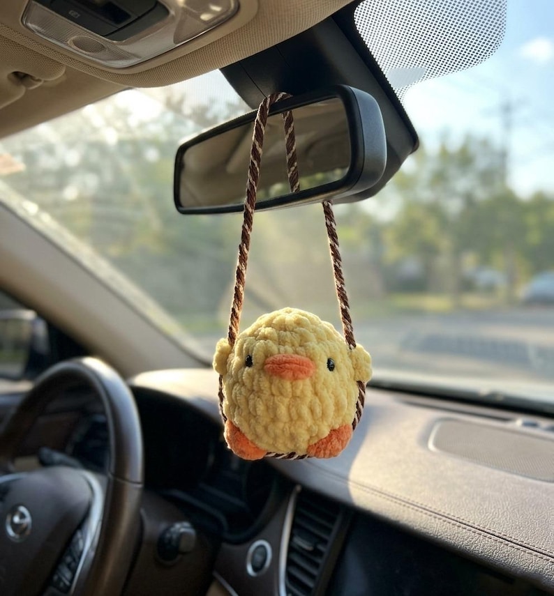 May include: A yellow crocheted chick car charm hanging from a car's rearview mirror. The chick has orange feet and a beak, with brown string straps. The interior of the car is visible, including the steering wheel and dashboard.
