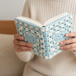 May include: A book held open, featuring a crocheted cover. The cover is a mix of light blue and cream-colored squares, with a heart-shaped design in the center. The book's pages are visible, and the cover is handmade.