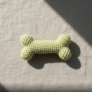 May include: A light green, crocheted dog bone toy. The toy is in the shape of a bone, with rounded ends and a textured surface. The toy is made of a soft, yarn-like material and is likely handmade. The toy is lying on a neutral-colored surface.
