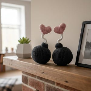 May include: Two handmade crochet sculptures. Each features a black sphere base, a white curly stem, and a pink heart-shaped top. They are displayed on a wooden shelf, with a framed picture and a potted plant in the background.