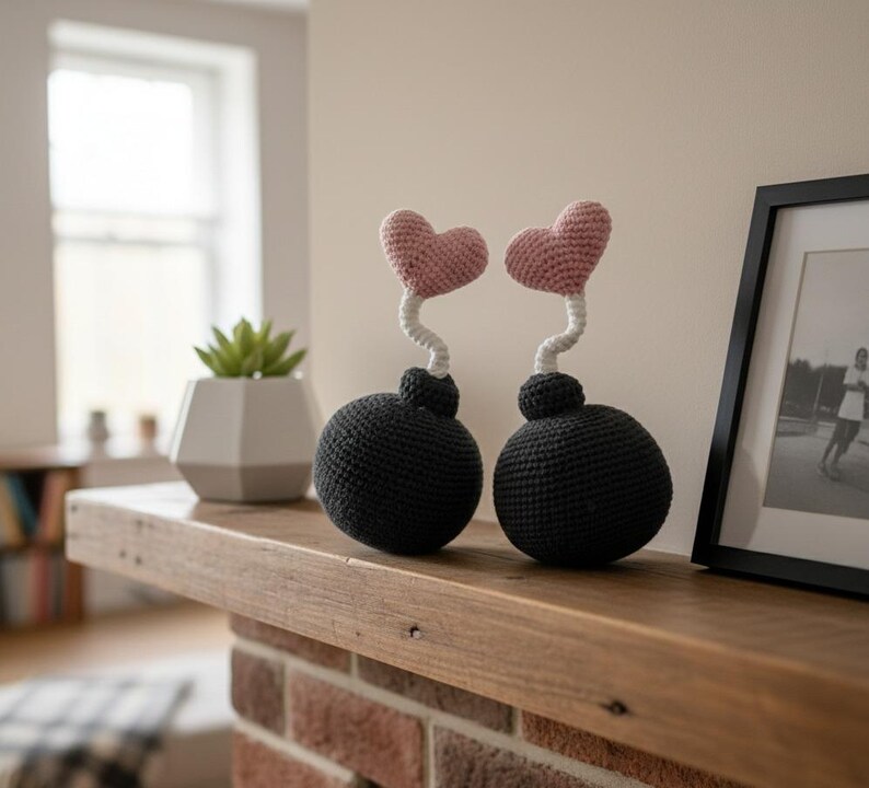 May include: Two handmade crochet sculptures. Each features a black sphere base, a white curly stem, and a pink heart-shaped top. They are displayed on a wooden shelf, with a framed picture and a potted plant in the background.