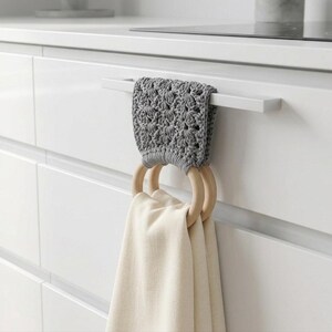 Peut inclure: Un porte-serviettes de cuisine en crochet gris avec deux anneaux en bois tenant une serviette crème. Le support est fixé à une poignée d'armoire blanche. L'arrière-plan montre une armoire de cuisine blanche.