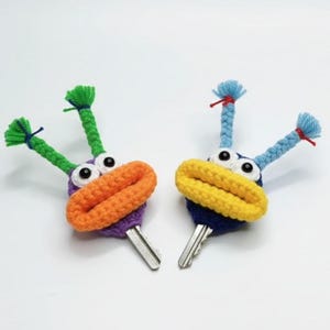 May include: Two whimsical key covers, each featuring a crocheted monster design. One has green antennae and an orange mouth, the other has blue antennae and a yellow mouth. Both have googly eyes and are attached to silver keys.