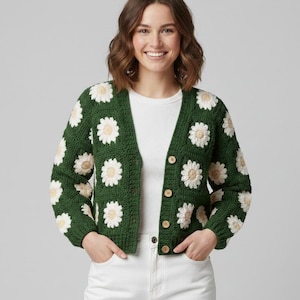 May include: A green crochet cardigan featuring daisy motifs. The cardigan has a V-neck, long sleeves, and wooden buttons. The daisies are white with a yellow centre. The person is wearing a white top and white trousers.