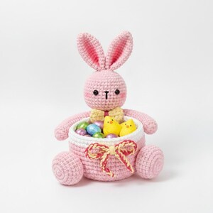May include: A pink crocheted Easter bunny holding a basket filled with colorful foil-wrapped eggs and yellow chick figures. The bunny has a yellow bow tie and a white and pink basket with a red and yellow bow.